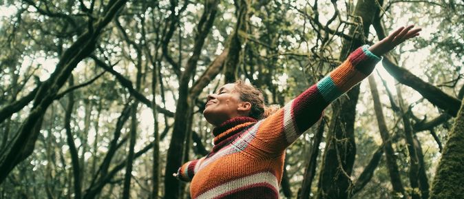 Eine Frau breitet glücklich die Arme aus und hat die Augen geschlossen, Sie steht in einem Wald voller dichter Bäume