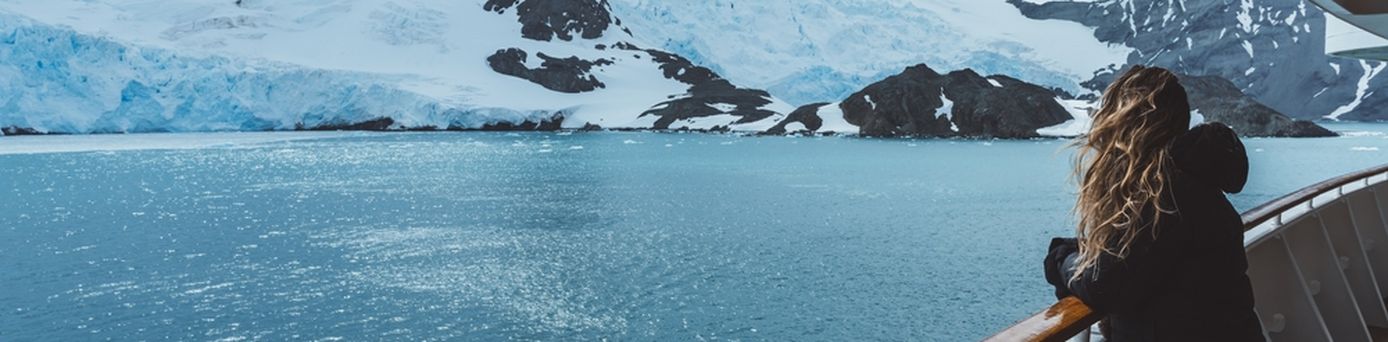 Eine Frau steht mit wehenden Haaren an der Reling eines Kreuzfahrtschiffes und schaut auf schneebedeckte Berge