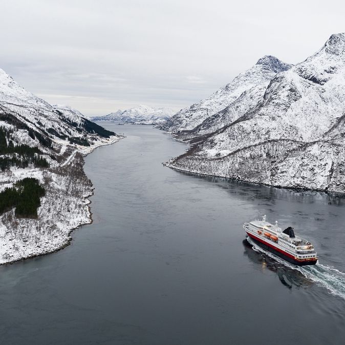 Ein Kreuzfahrtschiff fährt durch einen Fjord, umgeben von schneebedeckten Bergen und winterlicher Landschaft.