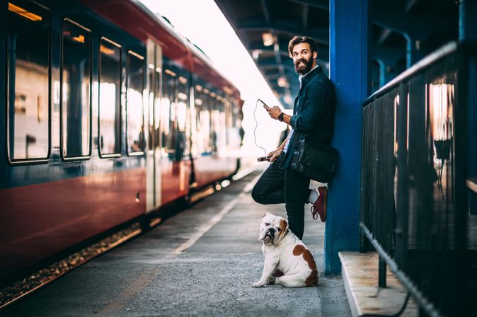 Ein Mann und ein Hund stehen gemeinsam an einem Bahnsteig und schauen in die Kamera