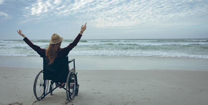 Frau im Rollstuhl hebt die Arme und schaut aufs Meer