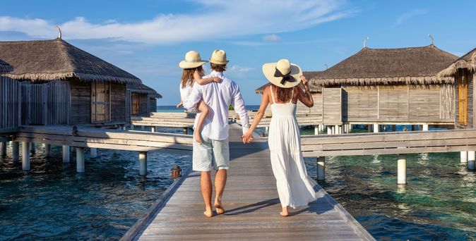 Eine Frau und ein Mann laufend Hand in Hand einen Steg auf dem Wasser entlang, der Mann trägt ein Kind auf dem Arm