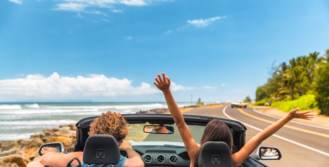 Zwei Personen im Auto mit offenem Verdeck, im Hintergrund kann man das Meer sehen