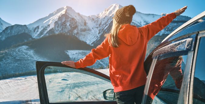 Eine Frau lehnt aus der offenen Autotür und schaut auf Schneebedeckte Berge
