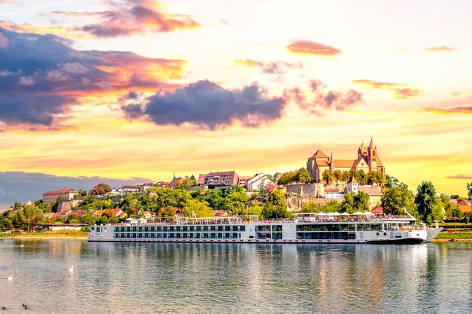 Langes Flusskreuzfahrschiff fährt entlang eines kleinen Dorfes, bei gelb leuchtendem Himmel
