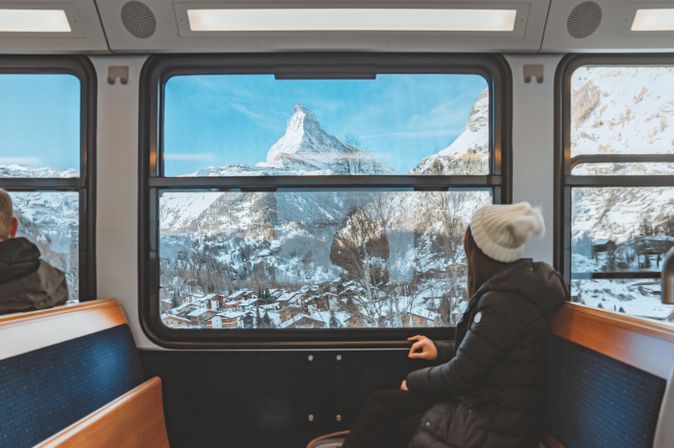 Eine Frau mit Winterjacke und Mütze sitzt in einem Zug und schaut aus dem Fenster in die Schneebedeckte Landschaft