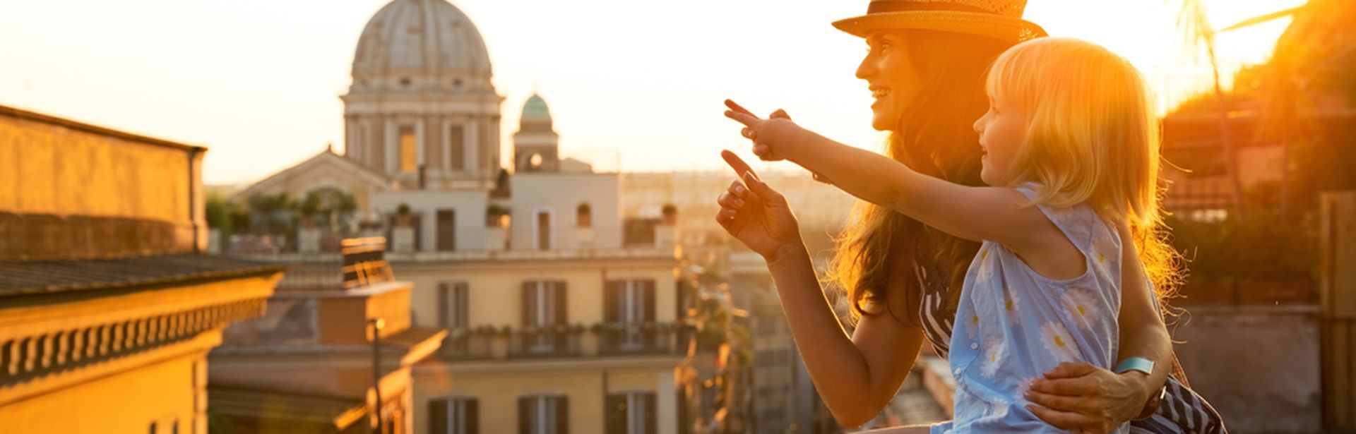 Eine Frau sitzt auf einer Mauer mit Ausblick auf eine Stadt. Auf ihrem Schoß sitzt ein kleines Mädchen und sie zeigen gemeinsam auf ein Bauwerk 