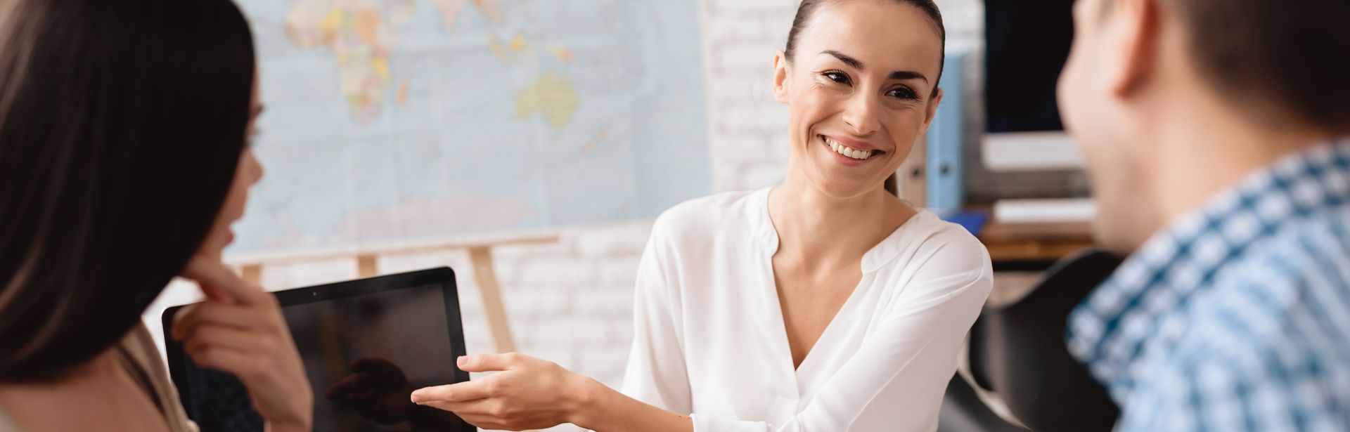 Ein Paar sitzt mit einer Reiseberaterin am Tisch, während die Reiseberaterin auf ein Tablet zeigt