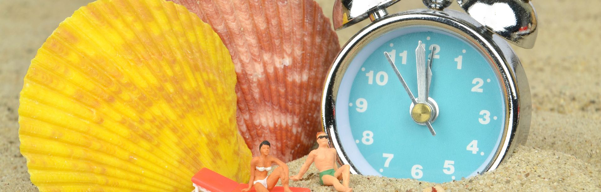 Im Sand liegen ein Wecker, zwei bunte Muscheln, ein Seestern und zwei Miniaturfiguren in Badekleidung