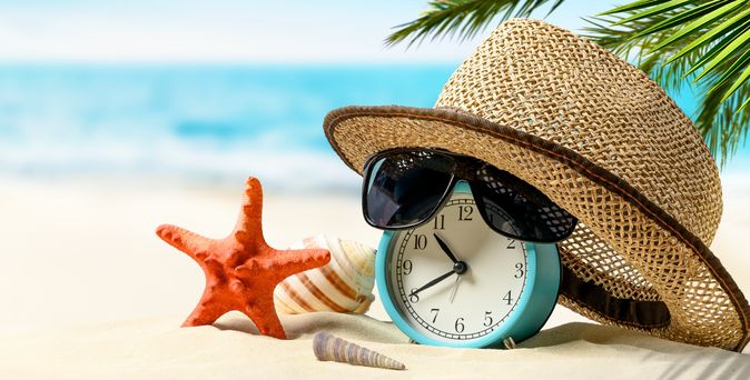 Ein Wecker mit Sonnenhut und einer Sonnenbrille liegen am Strand
