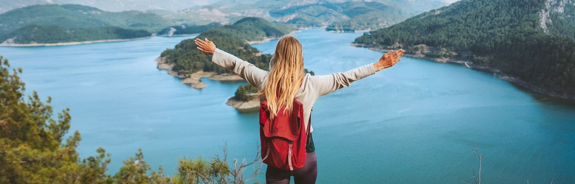 Eine Frau mit Rucksack schaut von einer Klippe auf einen großen See, eine Berglandschaft und breitet die Arme aus