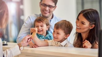 Eine Familie mit zwei Kindern beim Check-In in, Die Rezeptionistin reicht dem kleinen Kind die Zimmerkarte