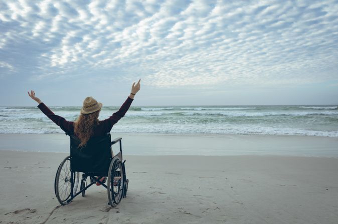 Frau im Rollstuhl hebt die Arme und schaut aufs Meer