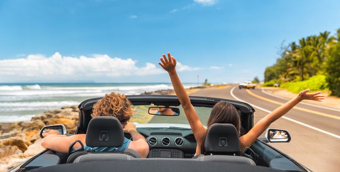 Zwei Personen im Auto mit offenem Verdeck, im Hintergrund kann man das Meer sehen