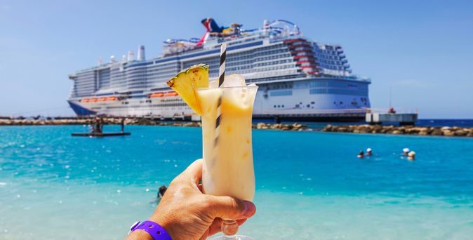 Eine Hand hält einen Cocktail hoch, im Hintergrund ist ein großes Kreuzfahrtschiff