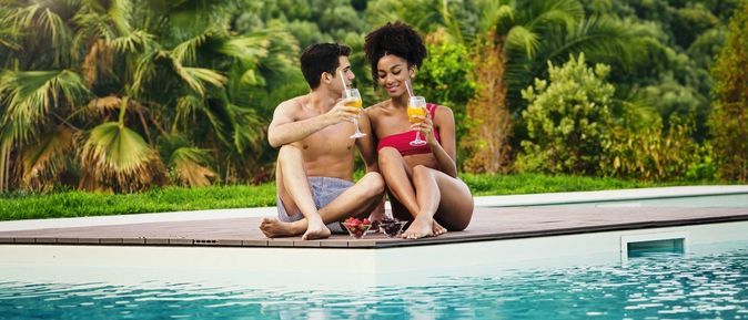 Paar sitzt mit Cocktails in der Hand und Obstschälchen auf der Terrasse vor einem Pool