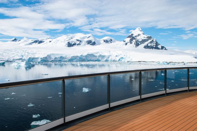 Ein Schiffsdeck mit Glasgeländer bietet einen Blick auf eine antarktische Landschaft mit schneebedeckten Bergen und schwimmenden Eisschollen