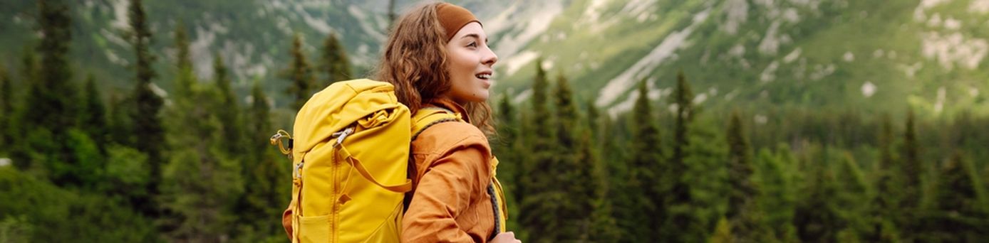 Eine Frau mit gelbem Wanderrucksack steht auf einem Berg und schaut begeistert in die Ferne