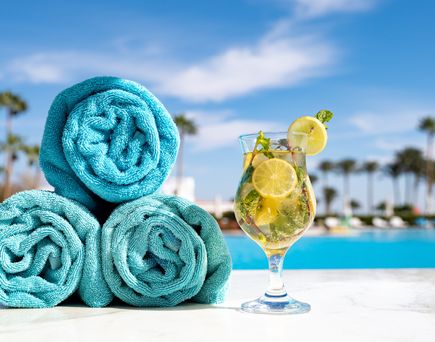 Drei aufgerollte Handtücher und ein Getränk stehen am Pool mit Palmen im Hintergrund