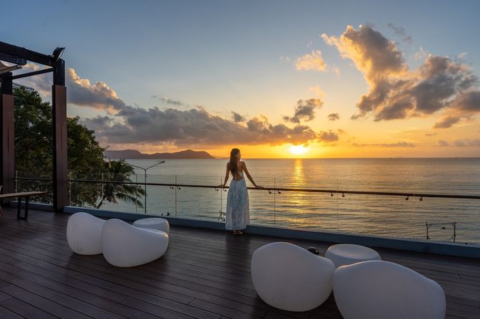 Frau am Geländer einer Terasse schaut auf den Sonnenuntergang über dem Meer