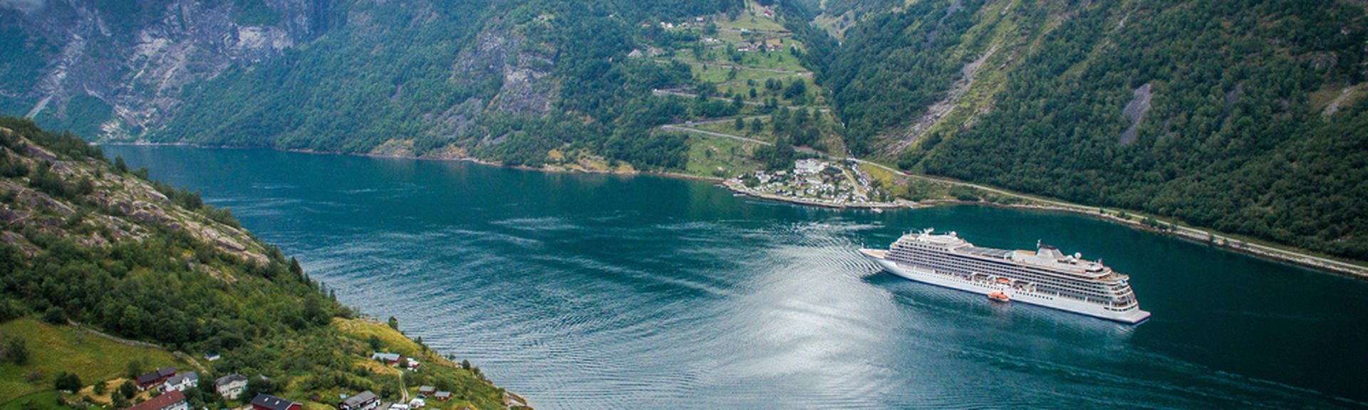 Ein Kreuzfahrtschiff fährt durch einen tiefen, von grünen Bergen gesäumten Fjord mit einem kleinen Dorf im Vordergrund.