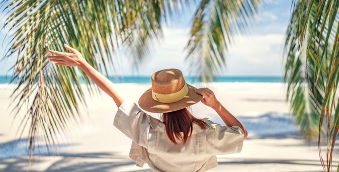 Frau mit Sonnenhut am Strand umgeben von Palmenblättern hebt einen Arm in die Luft