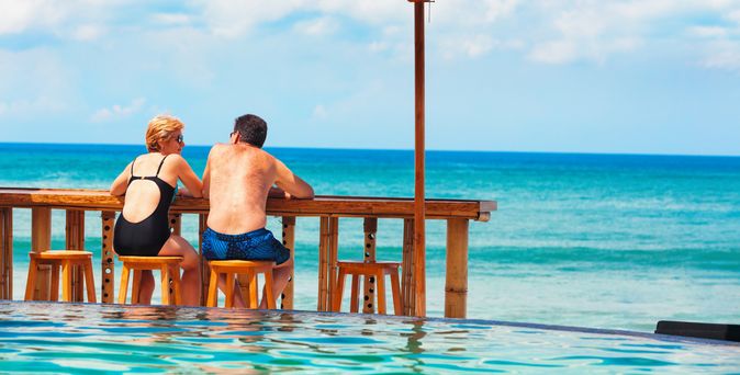 Eine Frau und ein Mann in Badekleidung sitzen an einer Holztheke auf Holzstühlen vor einem Pool und schauen auf das Meer