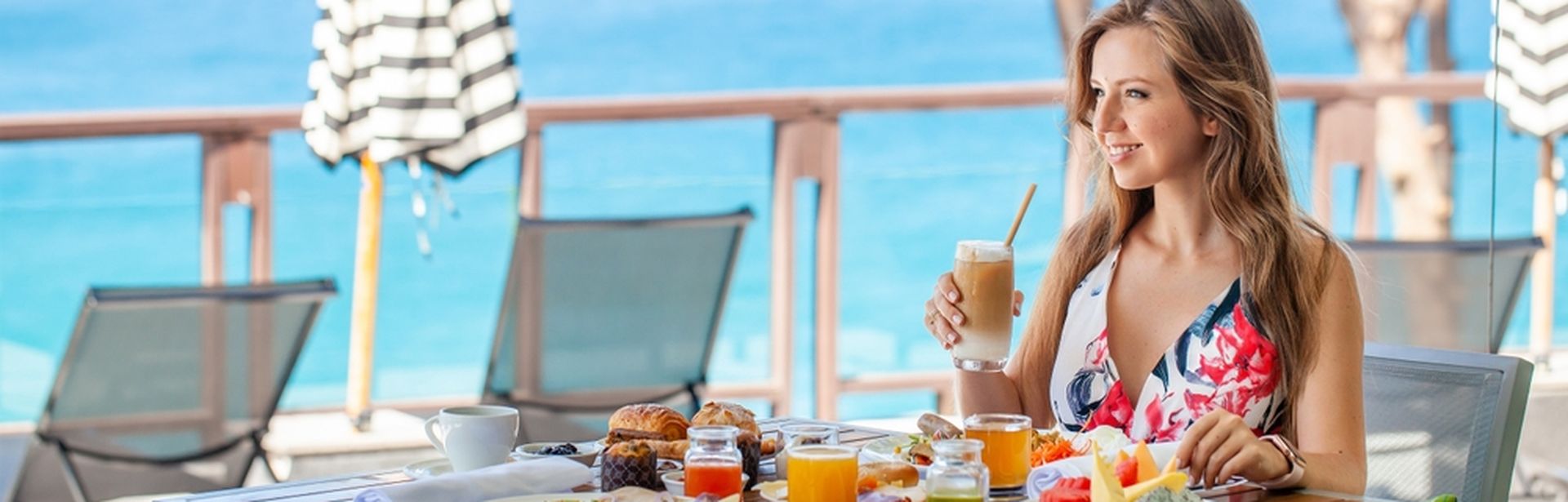 Frau sitzt lächelnd an einem gedeckten Tisch und hält einen Eiskaffee in der Hand