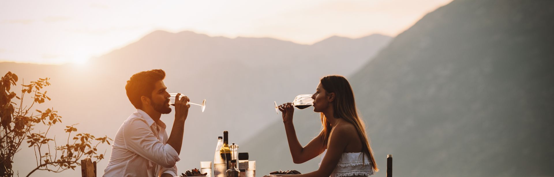 Eine Frau und ein Mann sitzen sich bei Sonnenuntergang an einem Tisch am Wasser gegenüber und trinken Wein