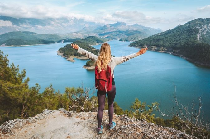 Eine Frau mit Rucksack schaut von einer Klippe auf einen großen See, eine Berglandschaft und breitet die Arme aus