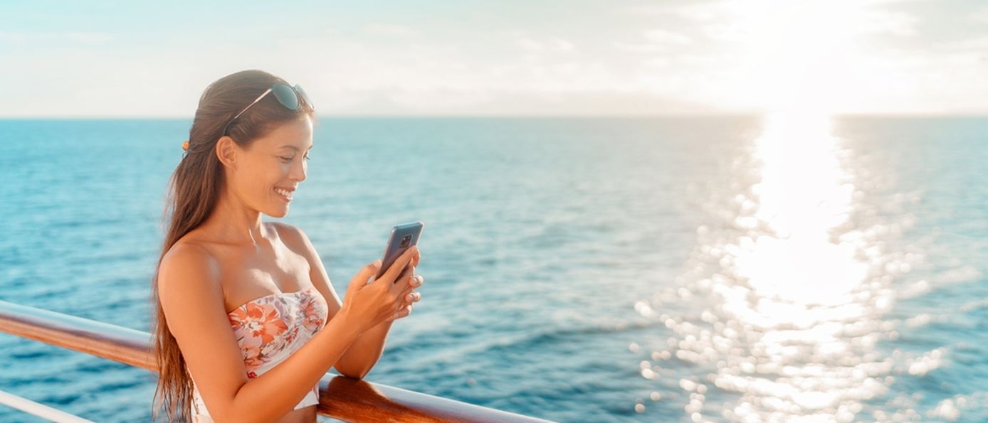 Eine Frau steht lächelnd an der Reling eines Kreuzfahrtschiffes und schaut auf ihr Smartphone