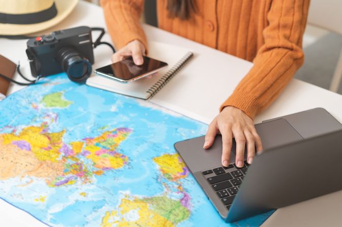Ein Schreibtisch mit einer Weltkarte als Unterlage, einem Sonnenhut und einer Kamera, eine Person bedient mit der einen Hand ein Handy und mit der anderen einen Laptop