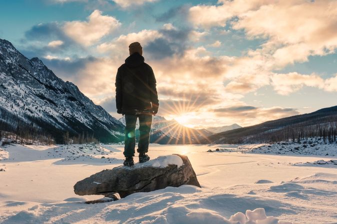 Mann steht leicht erhöht auf einem großen Stein in den Bergen bei Schnee und schaut in die untergehende Sonne