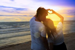 Ein Paar formt mit den Händen gemeinsam ein Herz und schaut bei Sonnenuntergang auf das Meer