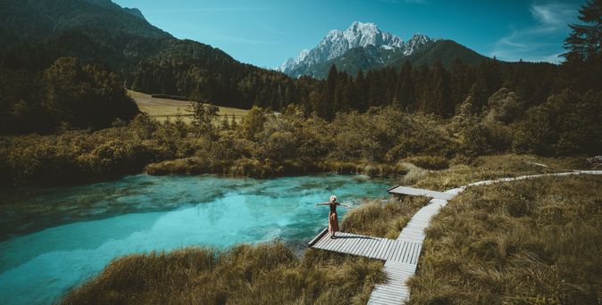 Eine Große Wiese mit einem Holzweg, auf einem Steg steht eine Frau und breitet die Arme aus, sie schaut auf den See vor ihr