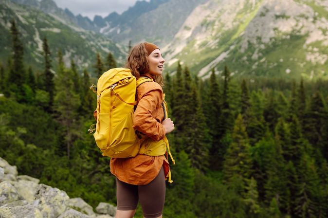 Eine Frau mit gelbem Wanderrucksack steht auf einem Berg und schaut begeistert in die Ferne