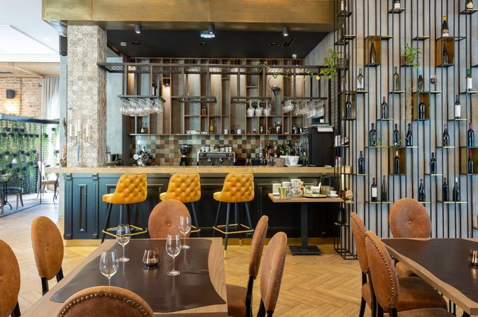 Eine Modern eingerichtete Bar mit gelben Barhockern und einer Wand mit Weinflaschen, im Vordergrund stehen mehrere mit Gläsern gedeckte Tische