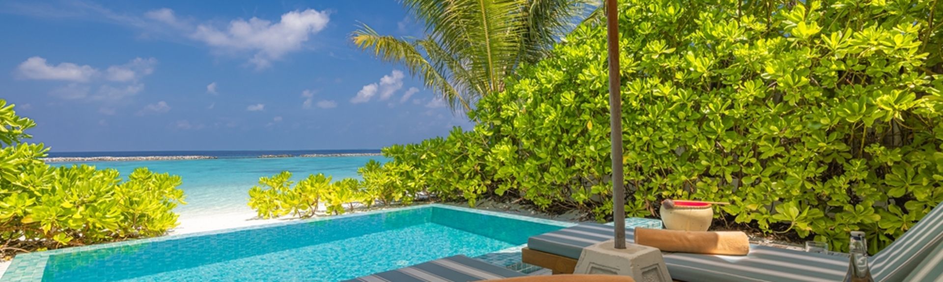 Zwei Liegen unter einem Sonnenschirm, davor ein kleiner Pool und der Strand 