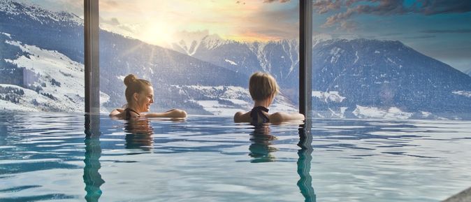 Zwei Frauen in einem Indoor Pool schauen aus dem Fenster auf schneebedeckte Berge