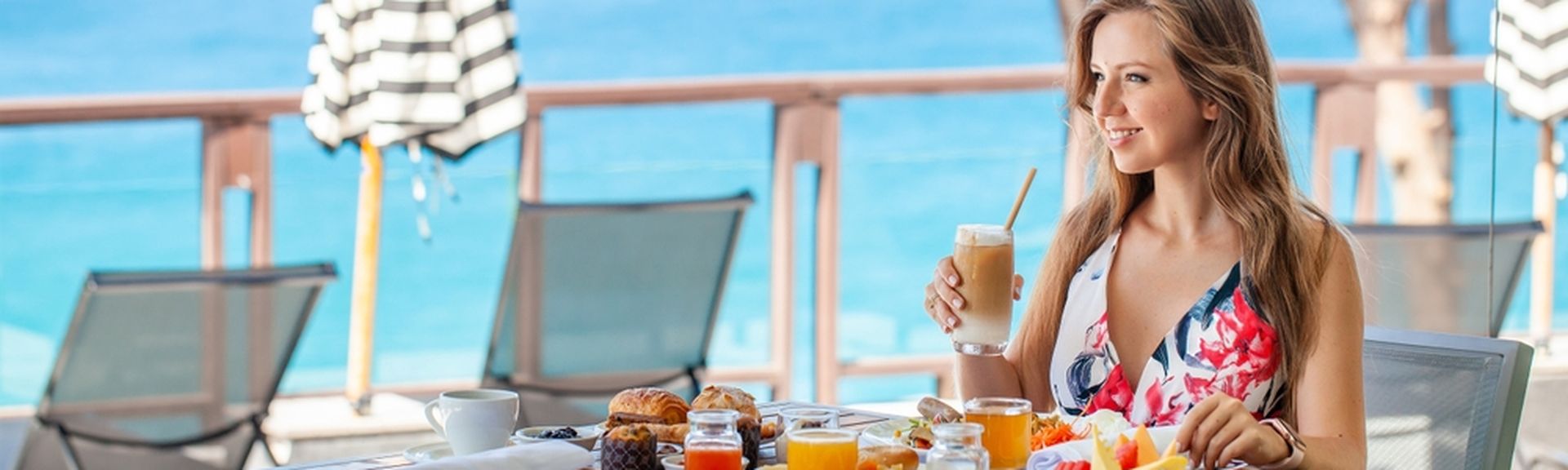 Frau sitzt lächelnd an einem gedeckten Tisch und hält einen Eiskaffee in der Hand