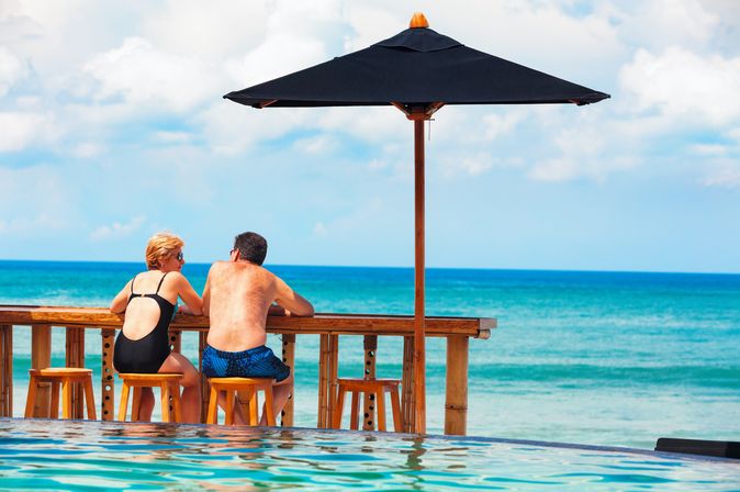 Eine Frau und ein Mann in Badekleidung sitzen an einer Holztheke auf Holzstühlen vor einem Pool und schauen auf das Meer