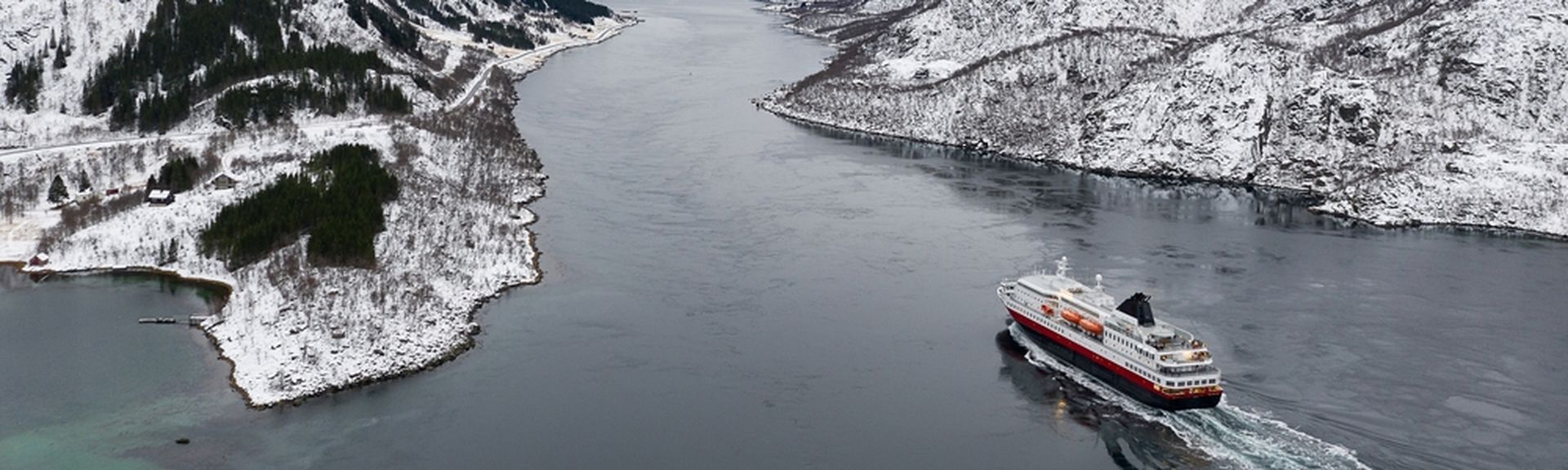 Ein Kreuzfahrtschiff fährt durch einen Fjord, umgeben von schneebedeckten Bergen und winterlicher Landschaft.