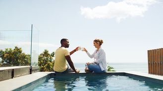 Eine Frau und ein Mann sitzen am Pool und stoßen mit Ihren Getränken an