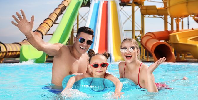 Eltern und ein Kind in einem Pool, Das Kind trägt eine Sonnenbrille und schwimmt in einem Schwimmring, im Hintergrund sind Wasserrutschen