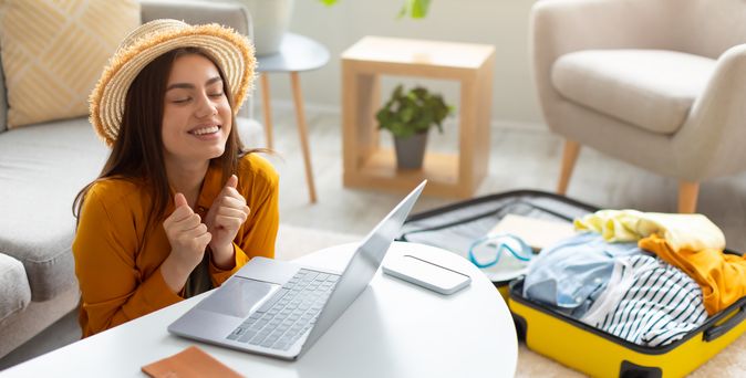 Eine sich freuende Frau sitzt vor einem Laptop. Im Hintergrund liegt ein gepackter Koffer