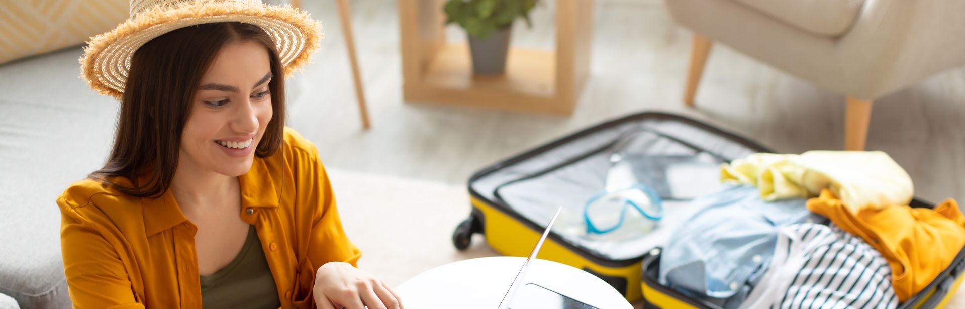 Eine sich freuende Frau tippt an einem Laptop. Im Hintergrund liegt ein gepackter Koffer