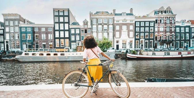 Eine Frau in einem gelben Rock steht an ihrem Fahrrad und schaut auf die Amstel und die gegenüberliegenden Häuser und Boote in Amsterdam