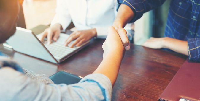 Zwei Personen geben sich über einem Schreibtisch die Hand und daneben sitzt eine weitere Person mit Laptop an dem Schreibtisch