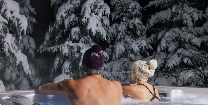 Ein Paar ist trägt Mützen, während sie im Whirlpool sind und nach außen zur Winterlandschaft blicken