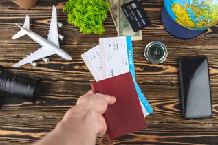 Auf einem Holztisch liegen ein Handy, ein Miniaturflugzeug, eine Kamera, ein Kompass, Geld und ein Globus. Eine Hand hält einen Reisepass und zwei Flugtickets
